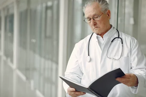 An older doctor wearing glasses and a stethoscope around his neck is standing and reading from a report in a black spiral-bound binder.