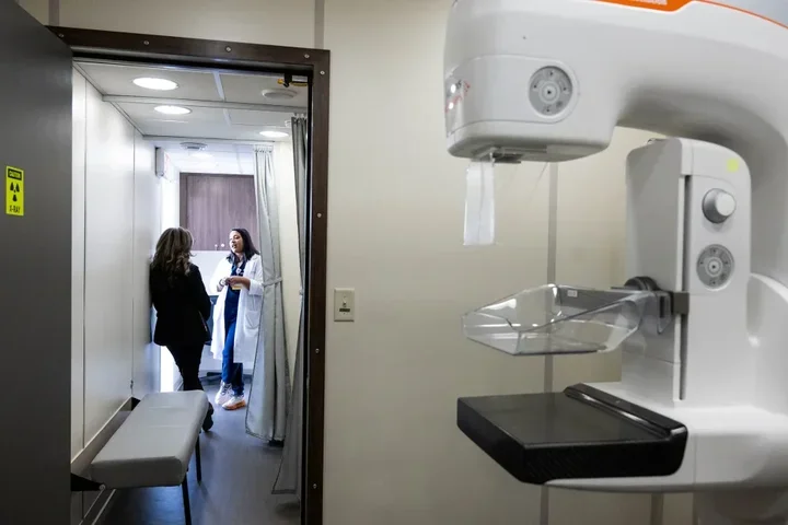 A mammography machine in the foreground at a clinic office with a doctor speaking with a patient in the background