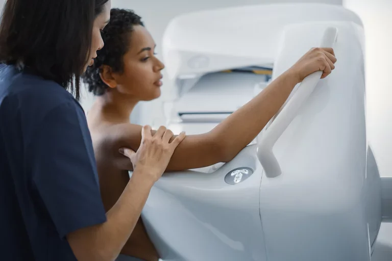 A woman stands against a mammogram machine. A mammographer stands behind her, positioning the first woman.