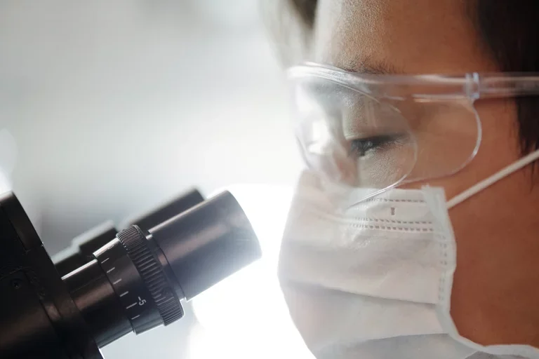 A researcher wearing protective glasses looks into a microscope.