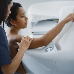 A woman stands against a mammogram machine. A mammographer stands behind her, positioning the first woman.