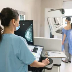A woman stands against a mammogram machine while a mammographer examines imaging.