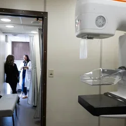A mammography machine in the foreground at a clinic office with a doctor speaking with a patient in the background
