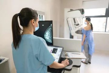 A woman stands against a mammogram machine while a mammographer examines imaging.